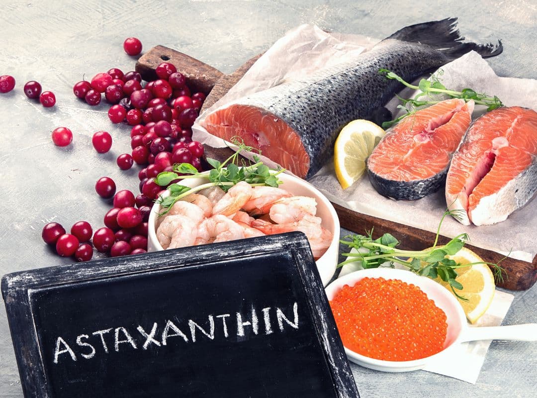 Auf dem Bild liegen frische Lebensmittel, die natürlicherweise Astaxanthin enthalten: Lachsfilets, Garnelen, roter Fischrogen und leuchtende Beeren. Im Vordergrund steht eine kleine Tafel mit der Aufschrift „Astaxanthin“.